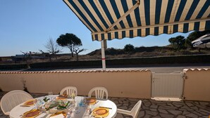 Un espace de restauration en plein air avec une table dressée pour quatre personnes, entourée de chaises en plastique blanches, sous une marquise à rayures.