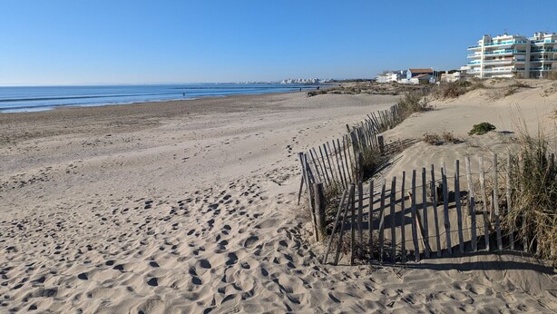 Une plage de sable avec des empreintes de pas, une clôture en bois et des bâtiments en arrière-plan.