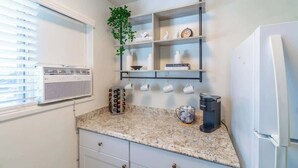 A kitchen with a granite countertop, white cabinets, a white refrigerator, a wall-mounted air conditioner, and a shelf with decorative items and a clock. - Emerald Escape - UNR & Pet Friendly - Fenced Yard (Reno)