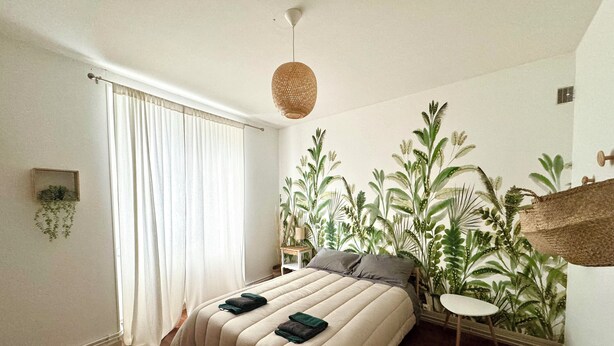 A bedroom with a bed, a hanging plant, a small table, and a window with curtains.