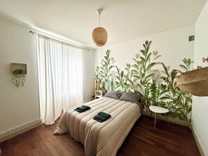 Un dormitorio con cama, una planta colgante, una mesita y una ventana con cortinas.