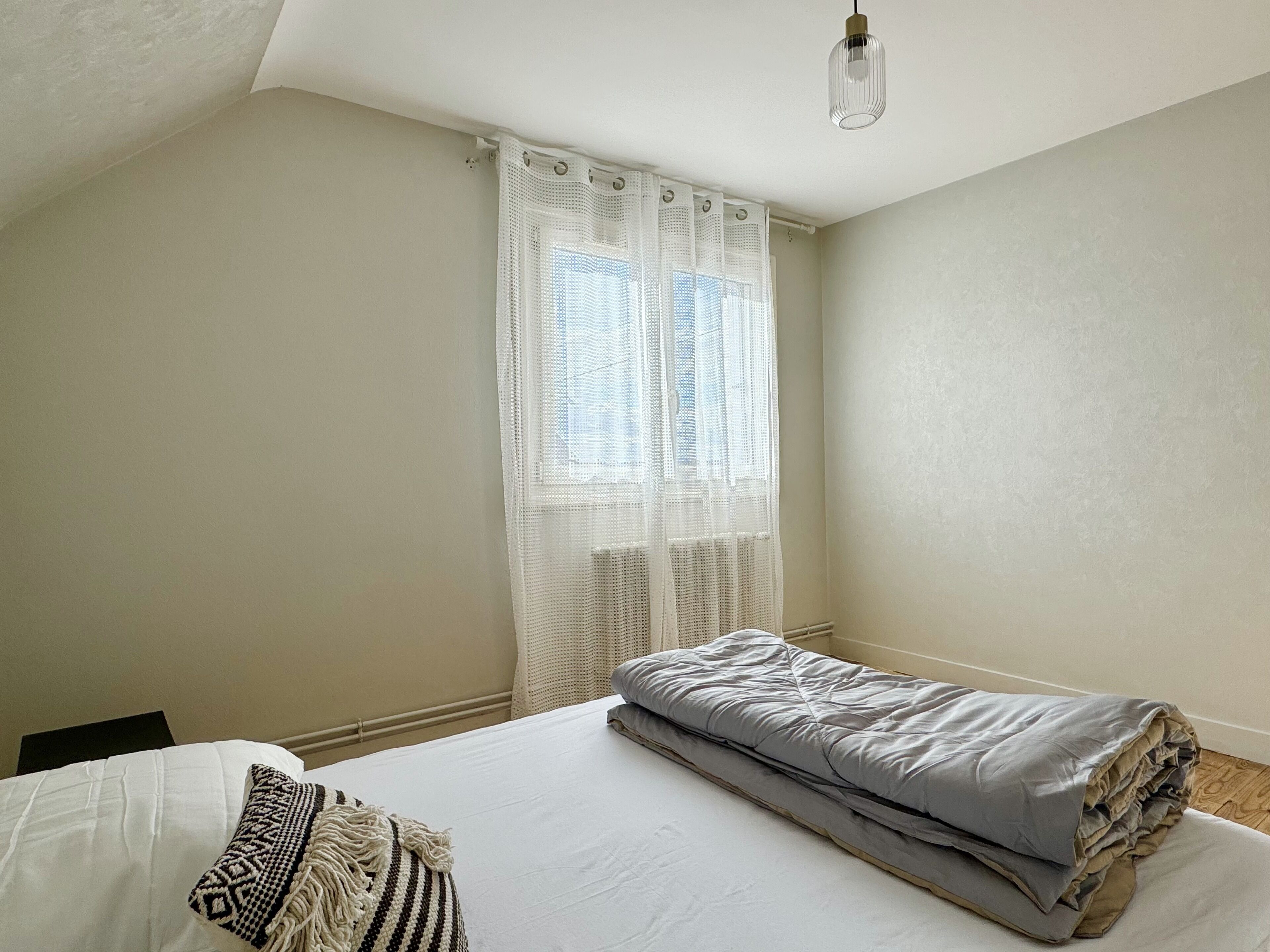A bedroom with a bed, a window with sheer curtains, and a hanging light fixture.