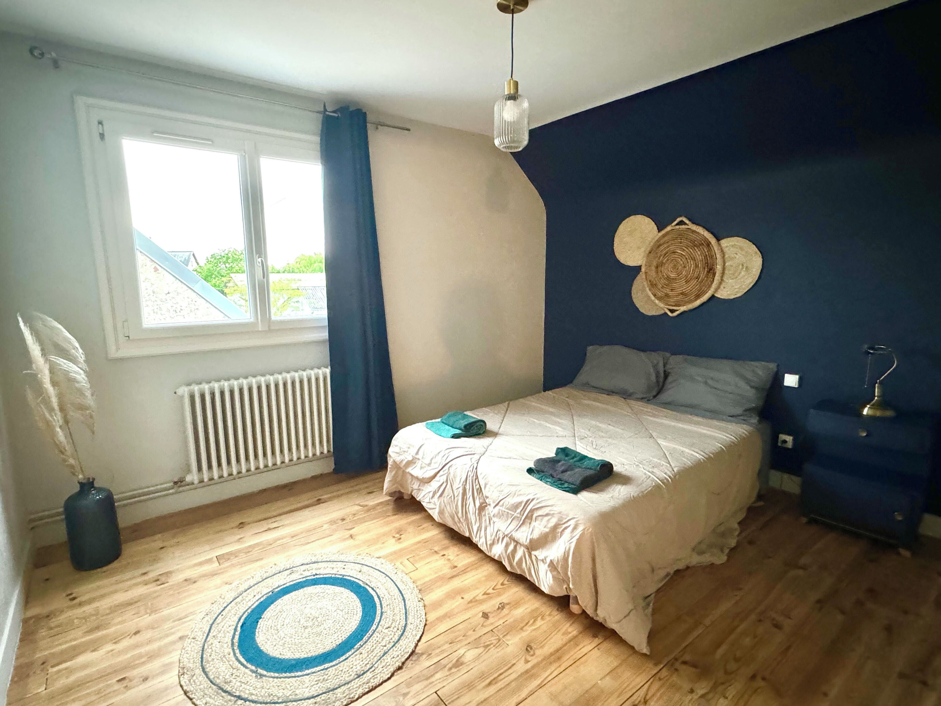 A bedroom with a bed, a radiator, a window with curtains, a blue wall with a decorative item, and a blue nightstand with a lamp.