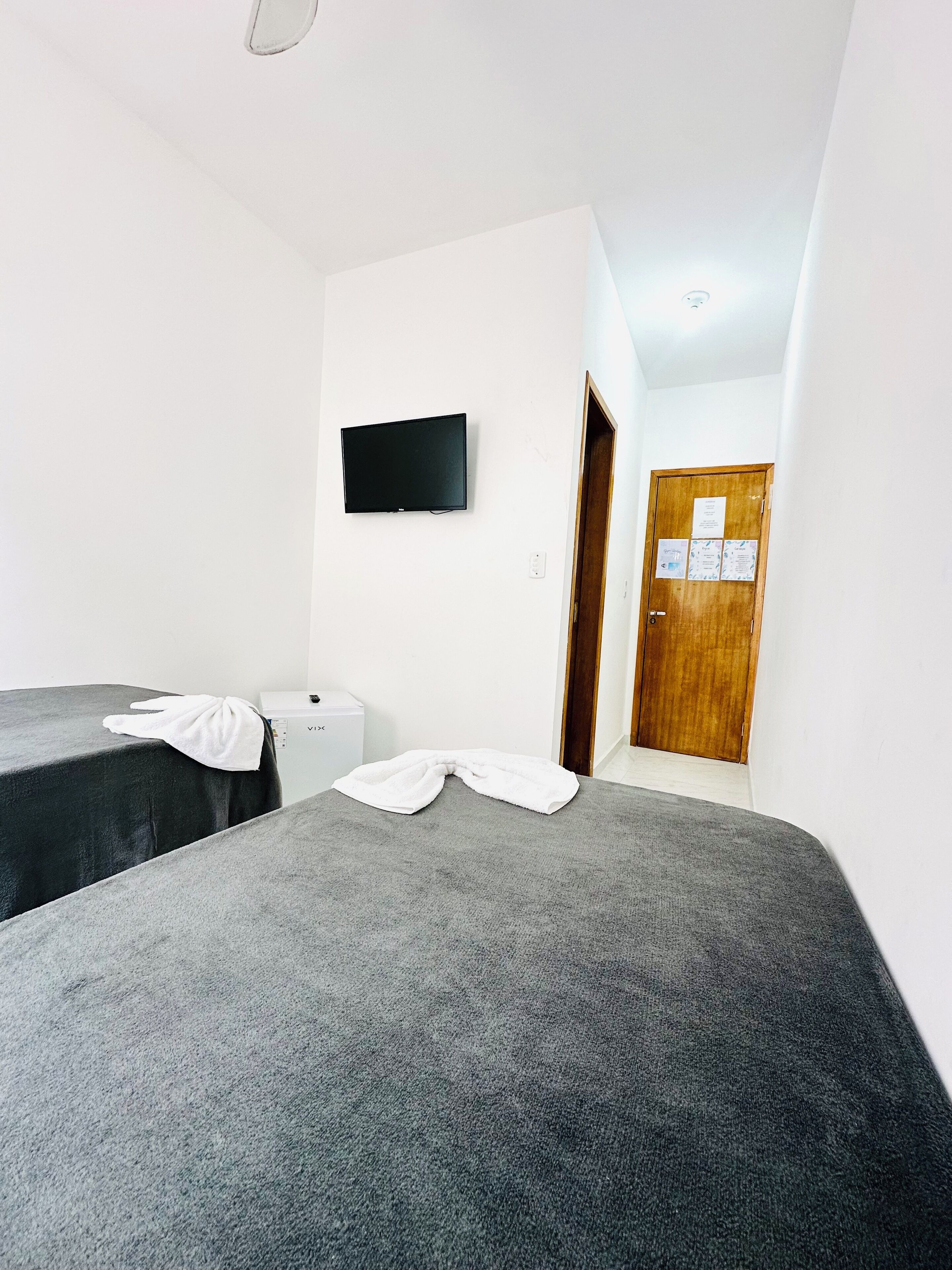 A hotel room with two beds, a TV mounted on the wall, a wooden door with a bulletin board, and a small white appliance.