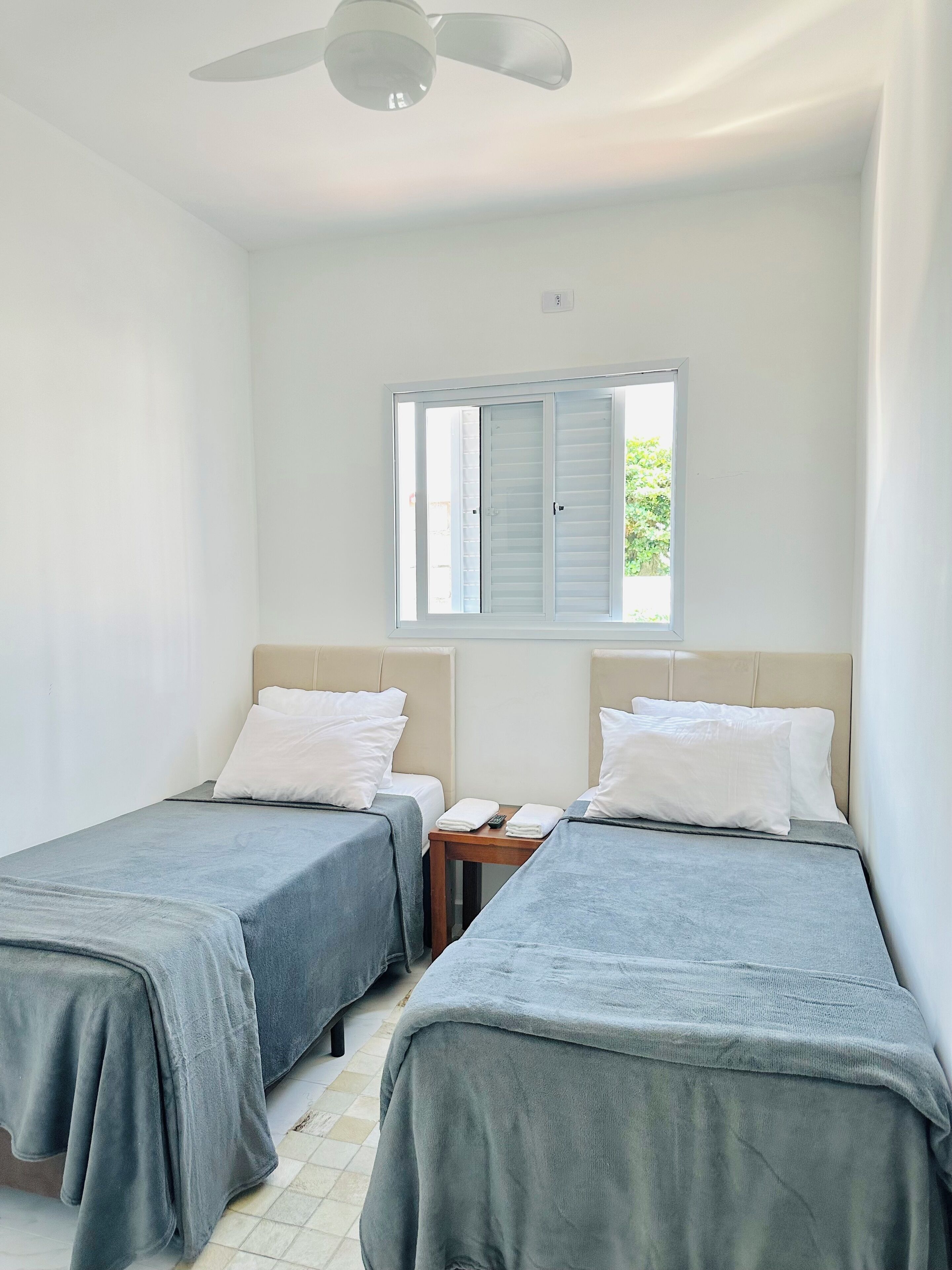 A hotel room with two single beds, a small table, a ceiling fan, and a window with shutters.