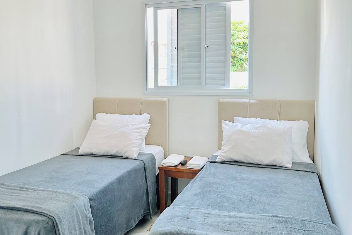 A hotel room with two single beds, a small table, a ceiling fan, and a window with shutters.