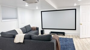 A modern living room with a gray sofa, a large flat-screen TV, and a ceiling projector. - Cozy and bright 6-bedroom house in Renton (Kent)