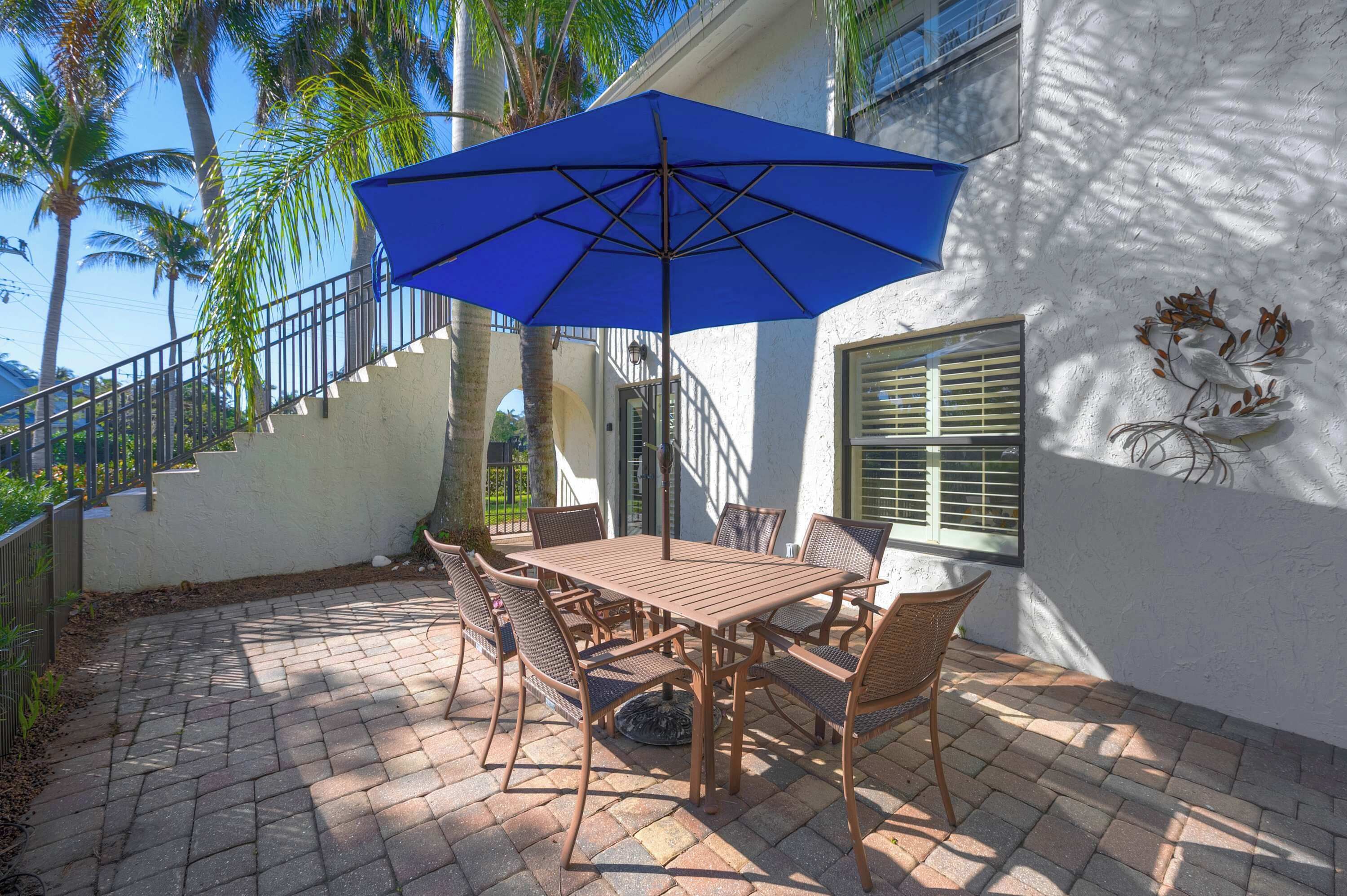 A patio with a wooden table and chairs, a blue umbrella, and a staircase leading to a balcony.