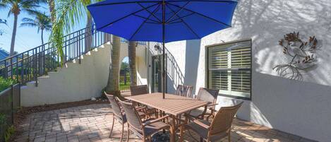 A patio with a wooden table and chairs, a blue umbrella, and a staircase leading to a balcony.