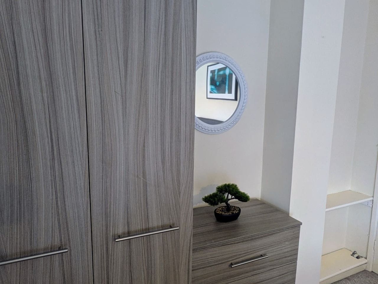 Ein Zimmer mit einem grauen Kleiderschrank, einer kleinen Holzeinrichtung mit Spiegel und einer Topfpflanze.