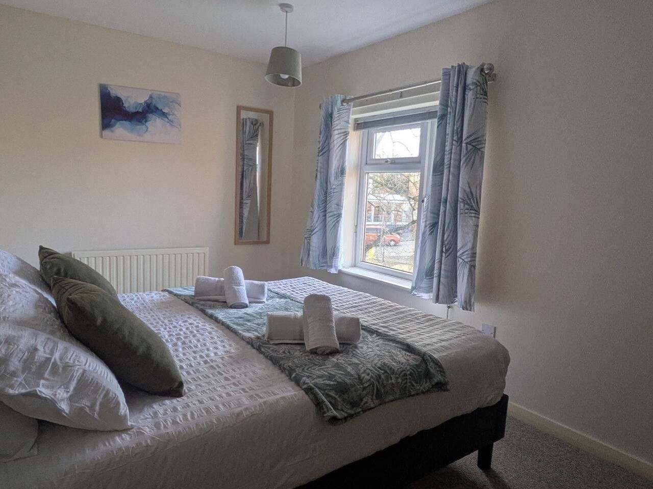 A neatly made bed with towels, a radiator, a window with curtains, and a painting on the wall.