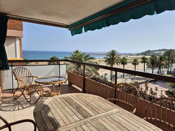 Un balcón con mesa de madera y sillas de mimbre, con vista a una playa con palmeras y cielo despejado.