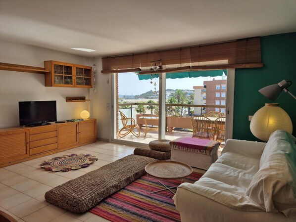 Een woonkamer met een televisie, een houten kast, een rieten bank en uitzicht op een balkon met buitenmeubilair.