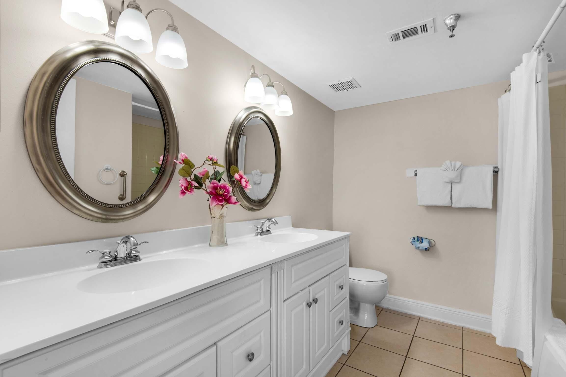 A bathroom with two sinks, a toilet, a shower curtain, and a mirror with lights.
