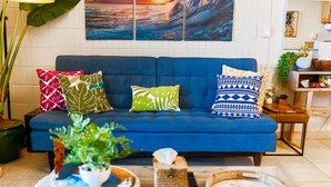 A living room with a blue sofa, decorative pillows, a large wall art of a wave, and a plant. - Oceanfront Hauula 1BR Condo I Beach, Pool, Parking (Hauula)