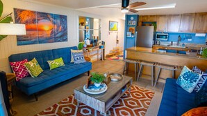 A modern living room with a blue sofa, a coffee table, and a kitchen area with wooden cabinets and a bar counter. - Oceanfront Hauula 1BR Condo I Beach, Pool, Parking (Hauula)