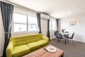Una habitación con un sofá verde, una mesa de centro de madera, una mesa de comedor con sillas y ventanas grandes con cortinas.