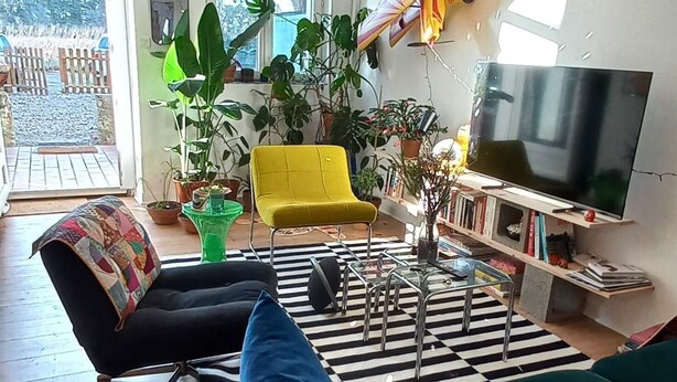 Una sala de estar moderna con un sofá verde, un sillón amarillo, un sillón negro y un tapete a rayas blanco y negro. Hay un televisor, una estantería y un ventanal con vistas al exterior.