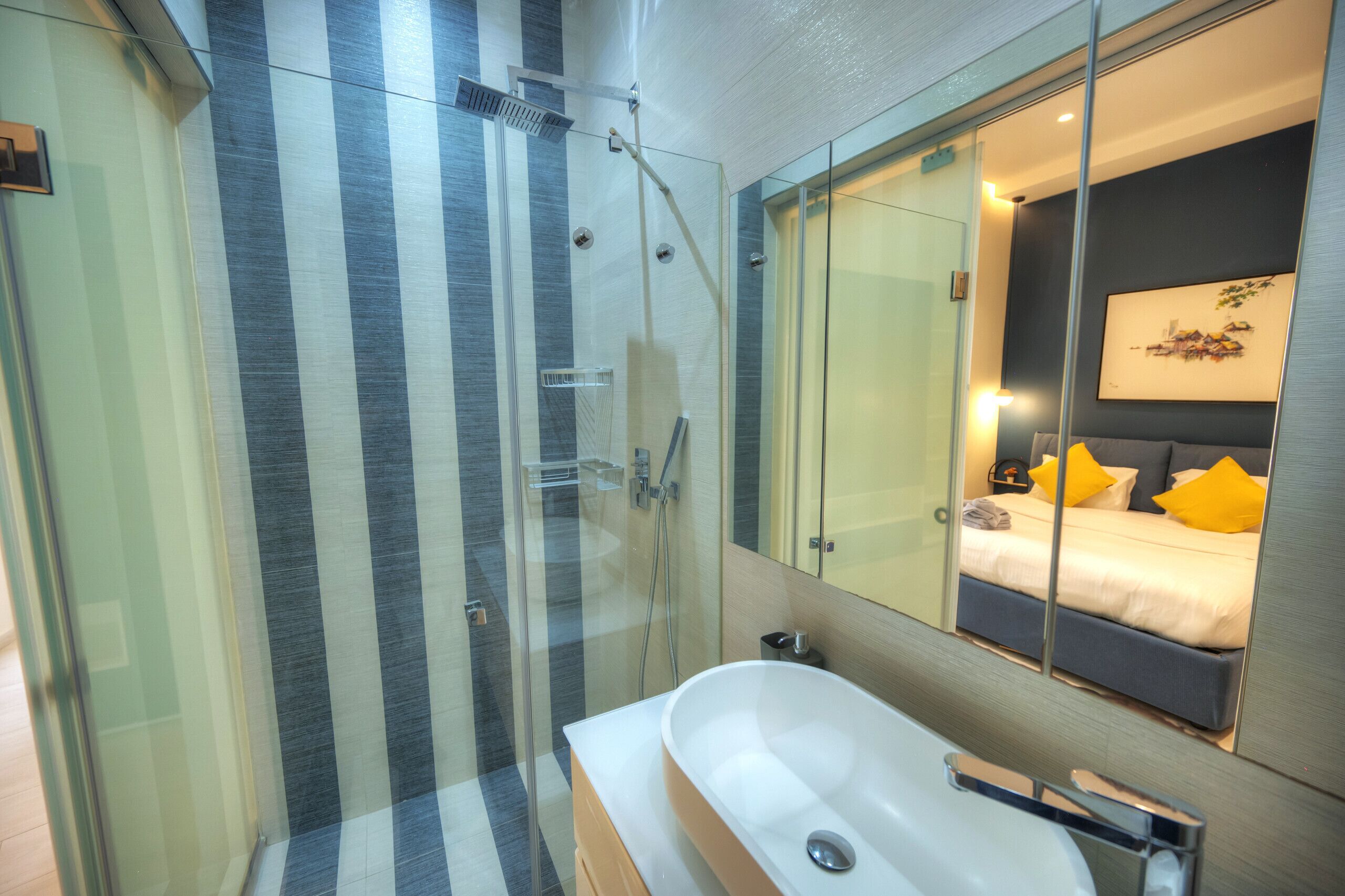 Ein modernes Badezimmer mit einer Duschkabine aus Glas, einer Badewanne und einem großen Spiegel, der ein Bett mit gelben Kissen reflektiert.