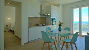 A modern kitchen with white cabinets, a stainless steel range hood, and a built-in oven. There is a dining area with a round table and four chairs. A vase with flowers is on the table. - IRIS Aurora Apartments (Baia Azzurra-levagnole)