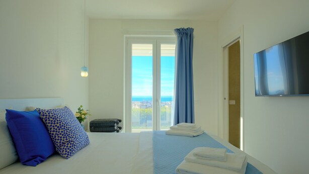 A hotel room with a bed, a TV, a view of the sea, and a suitcase on the floor. - IRIS Aurora Apartments (Baia Azzurra-levagnole)