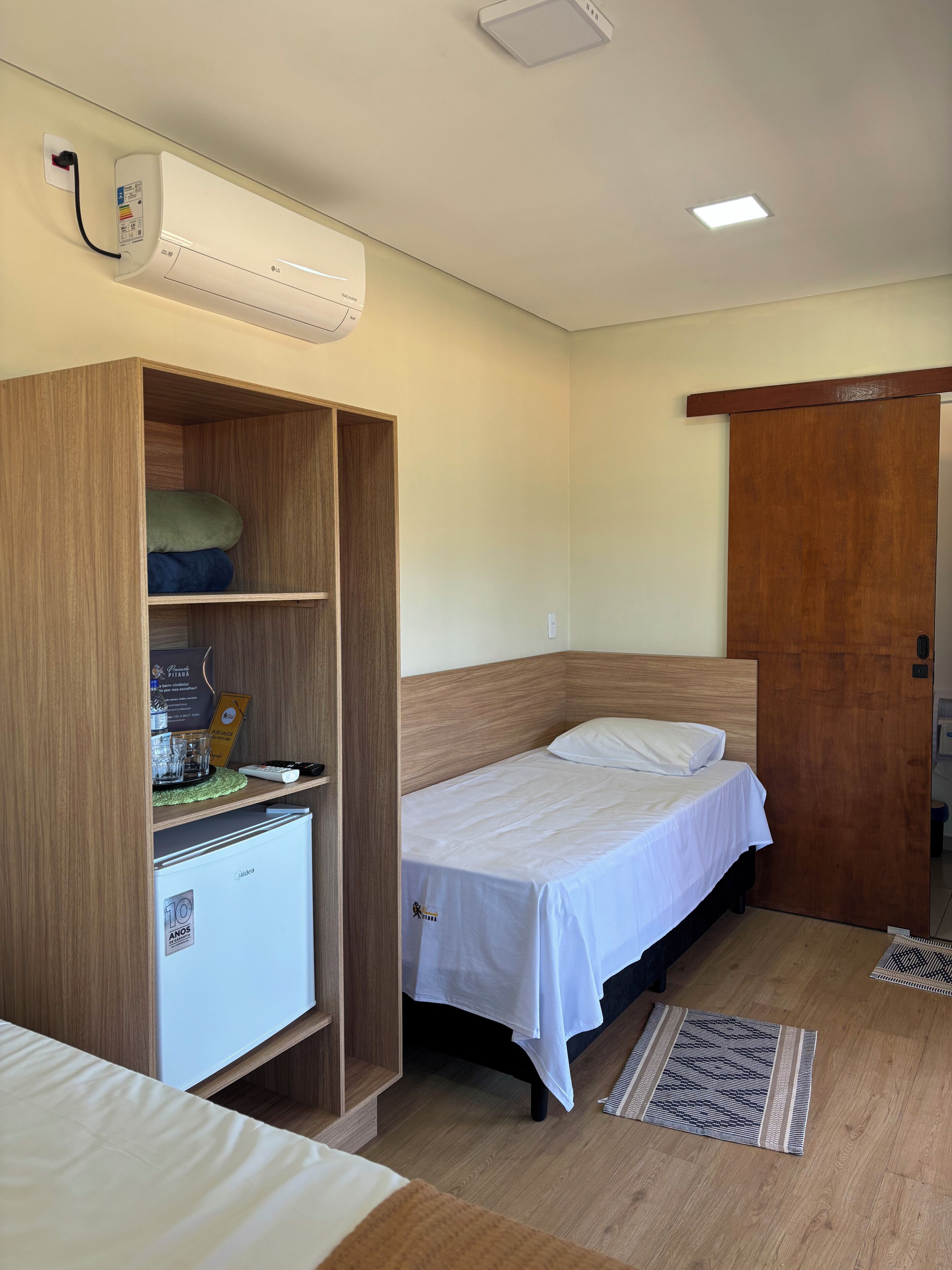 A compact room with a bed, a wardrobe, a small refrigerator, and a ceiling-mounted air conditioner.