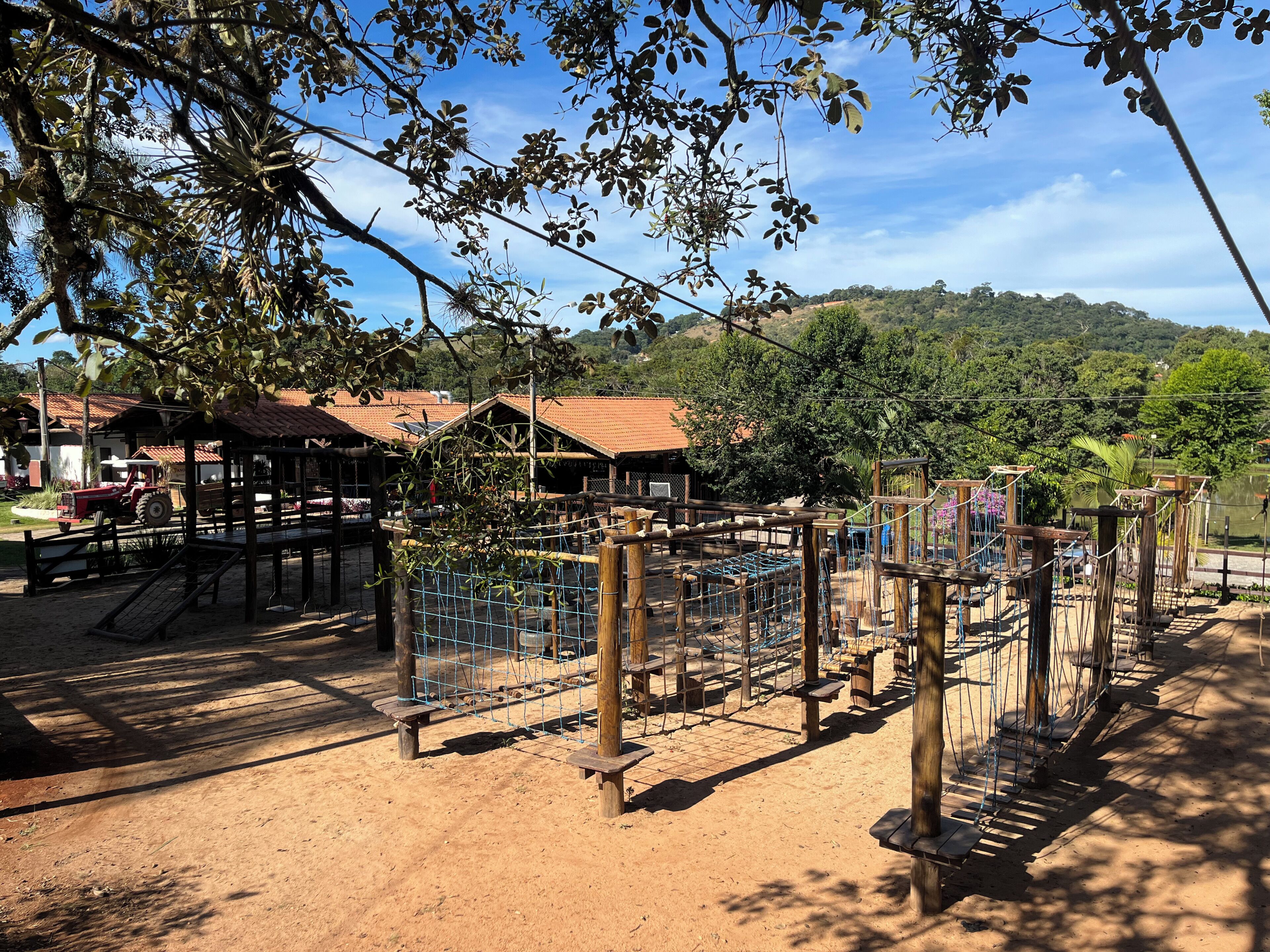 Un parque infantil con estructuras de madera y puentes de cuerda, rodeado de árboles y suelo de tierra.