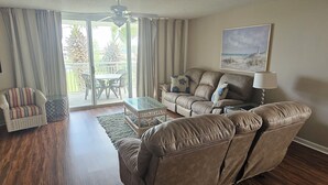 Un salon comprenant un canapé, un fauteuil et une table basse en verre. Il y a un balcon avec une table et des chaises à l’extérieur.