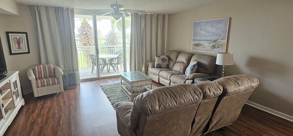 A living room with a sofa, armchair, and a glass coffee table. There is a balcony with a table and chairs outside.