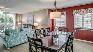 Une chambre d’hôtel dotée d’un coin repas comprenant une table et des chaises en verre, un canapé à motifs étoilés et une grande fenêtre avec des stores.