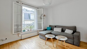 A modern living room with a grey sofa, two small side tables, and a large window with a view of buildings and plants outside. - Cozy apartment in the 11th arrondissement (Paris)