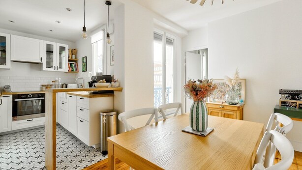 A modern kitchen with white cabinets, a wooden dining table, and a vase of dried flowers. - Cozy apartment in the 11th arrondissement (Paris)