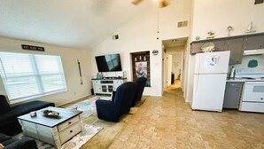 A living room with a TV, a sofa, a coffee table, and a kitchen area with a refrigerator and stove. - Fishing Haven at Linda's Mermaid Cove (Freeport)