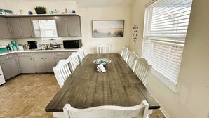 A dining area with a wooden table and white chairs, a kitchen with grey cabinets, and a window with blinds. - Fishing Haven at Linda's Mermaid Cove (Freeport)