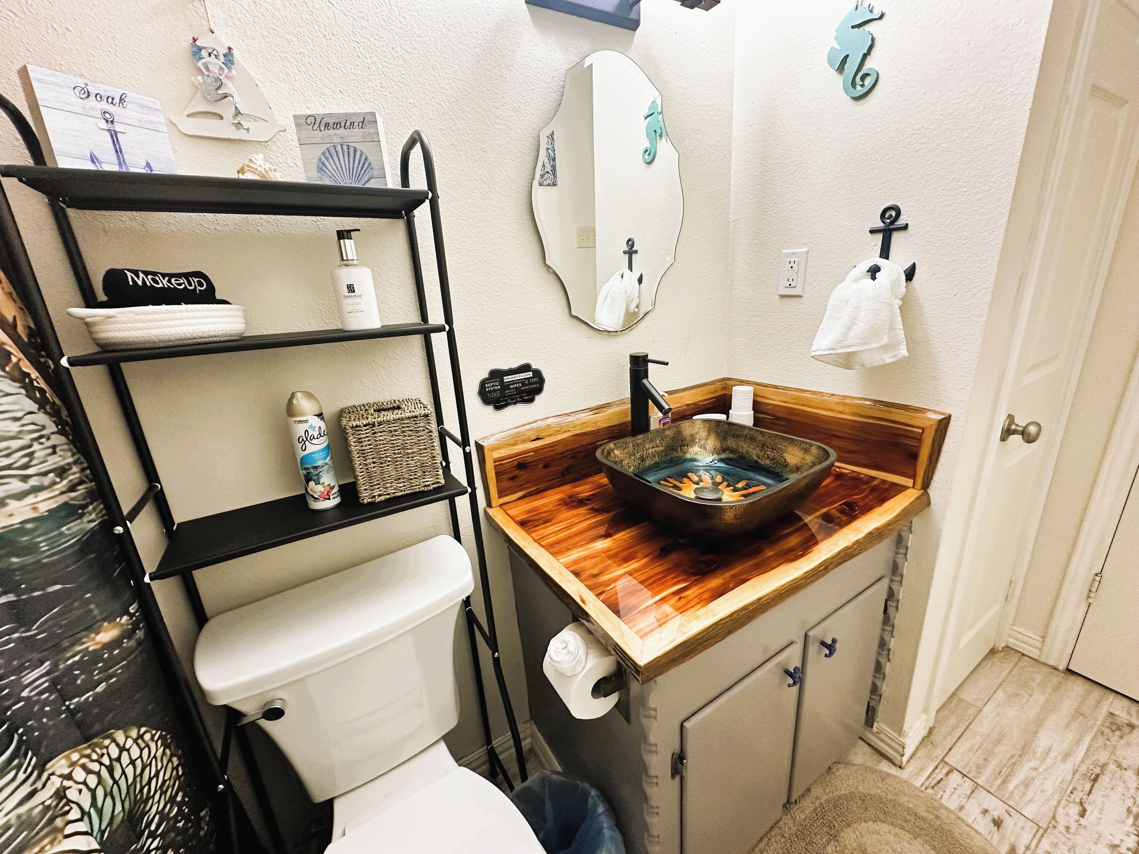 Un baño con tocador de madera, estante metálico negro con artículos de aseo, inodoro blanco y espejo con marco decorativo.