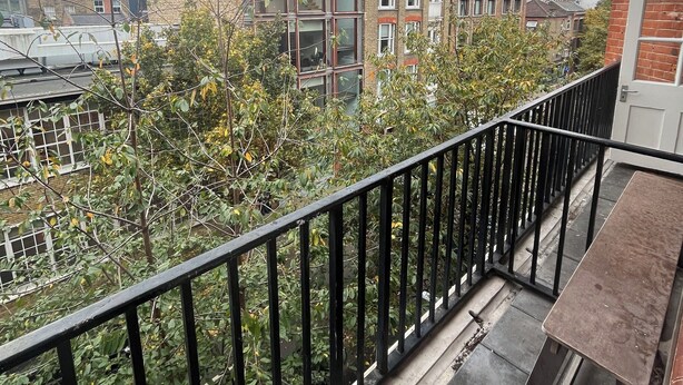 A balcony with a black metal railing, a small table, and a view of surrounding buildings and trees.