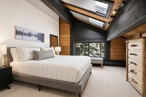 A bedroom with a large bed, a wooden dresser, a bench, and a window with a view of snow-covered trees.