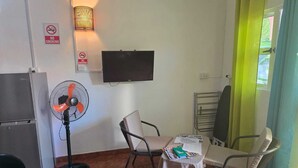 Een kleine kamer met een televisie, een ventilator, een tafel met een stoel en een raam met gordijnen.