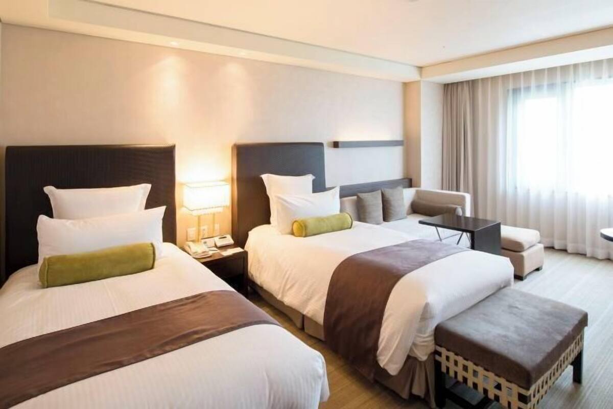 Ein Hotelzimmer mit zwei Betten, einem Sofa, einem kleinen Tisch und einem Fenster mit Vorhängen.