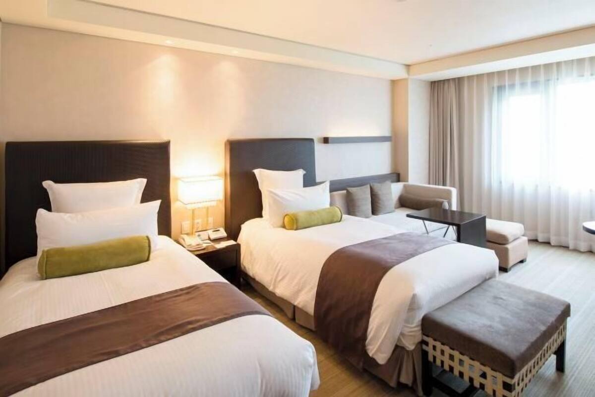 Ein Hotelzimmer mit zwei Betten, einem Schreibtisch, einem Stuhl und einem fensterbepflanzten Fenster.