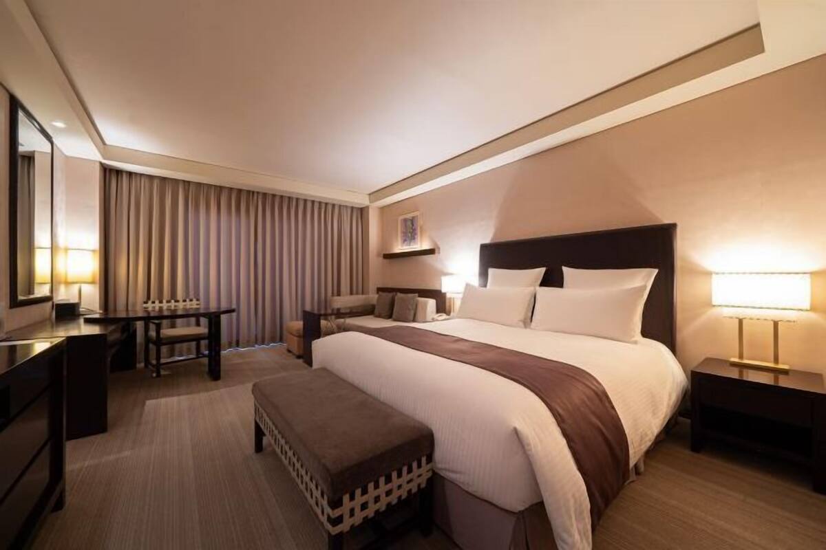 Ein Hotelzimmer mit einem großen Bett, Nachttischen, einem Schreibtisch, einem Stuhl, einer Bank und einem Spiegel.