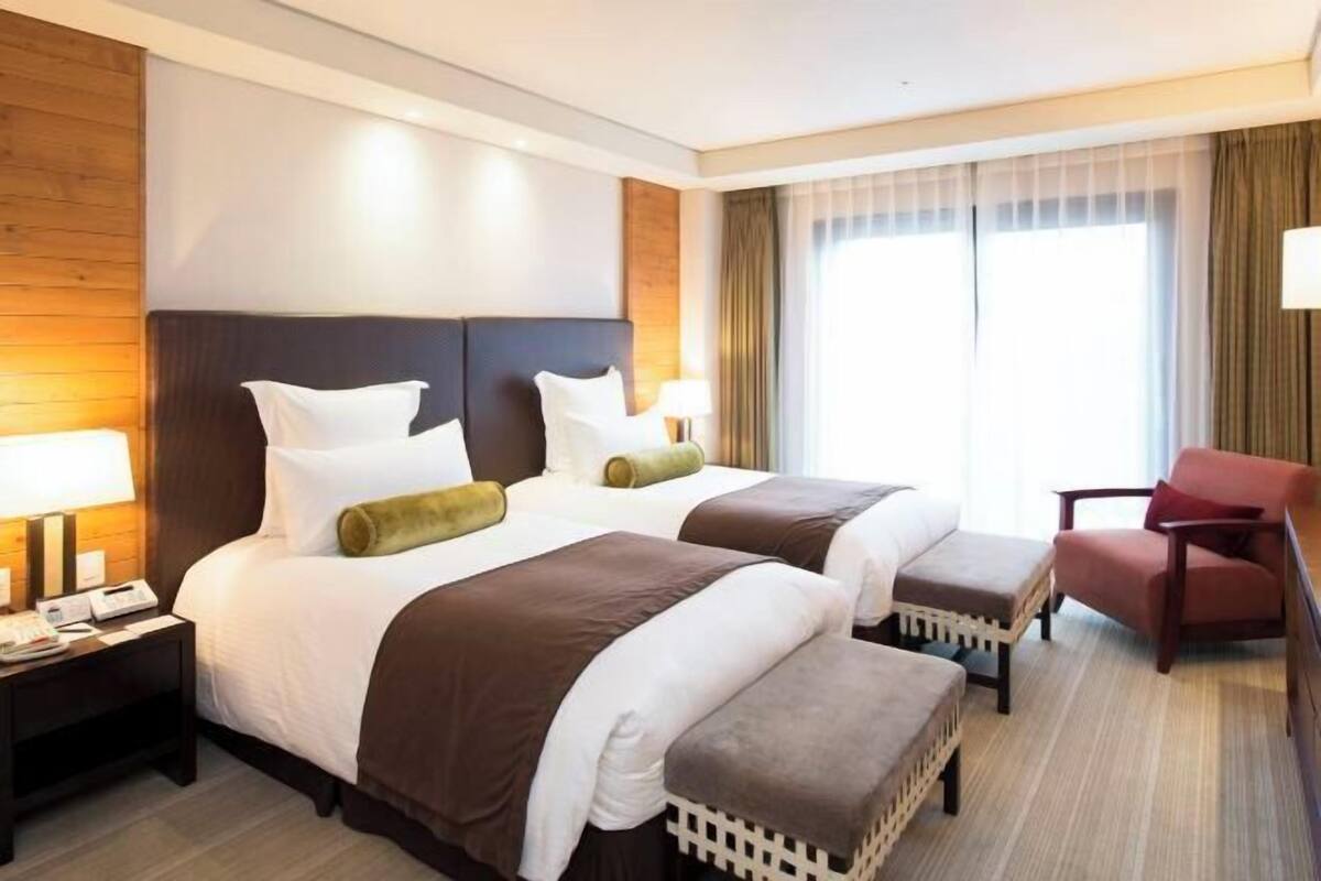 Ein Hotelzimmer mit zwei Betten, einem Nachttisch, einem Sessel und einem großen Fenster mit Vorhängen.
