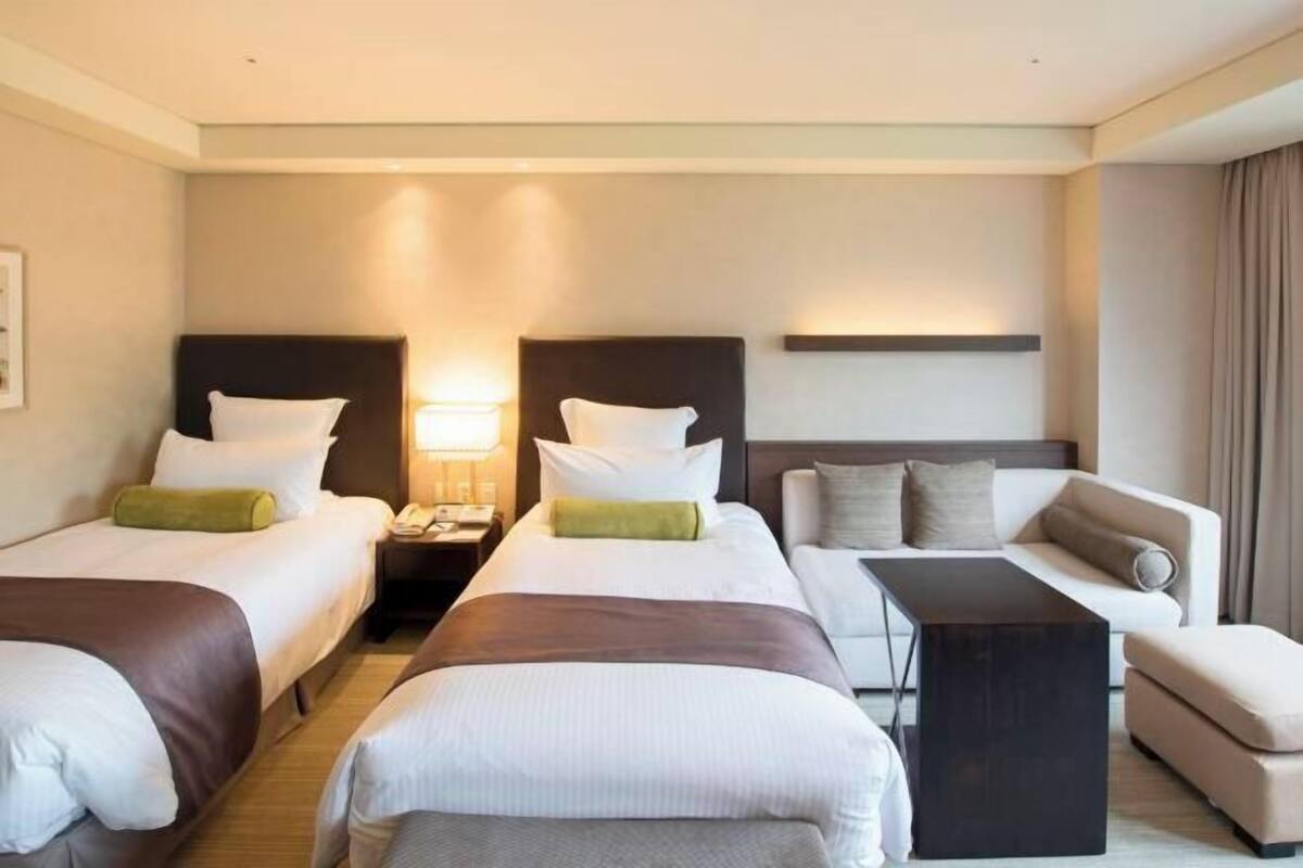 Ein Hotelzimmer mit zwei Einzelbetten, einem Schlafsofa, einem kleinen Tisch und einem Nachttisch mit Lampe.