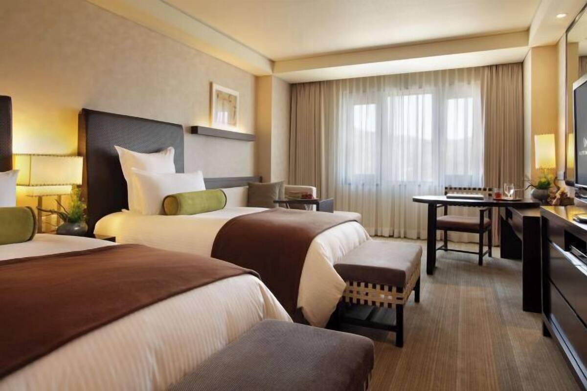 Ein Hotelzimmer mit zwei Betten, einem Schreibtisch, einem Fernseher und einem großen Fenster mit Vorhängen.