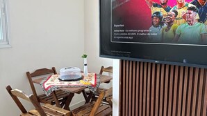 A dining area with a table and chairs, a television mounted on a wall, and a wooden panel. - CASA DOS VENTOS (Praia Grande)