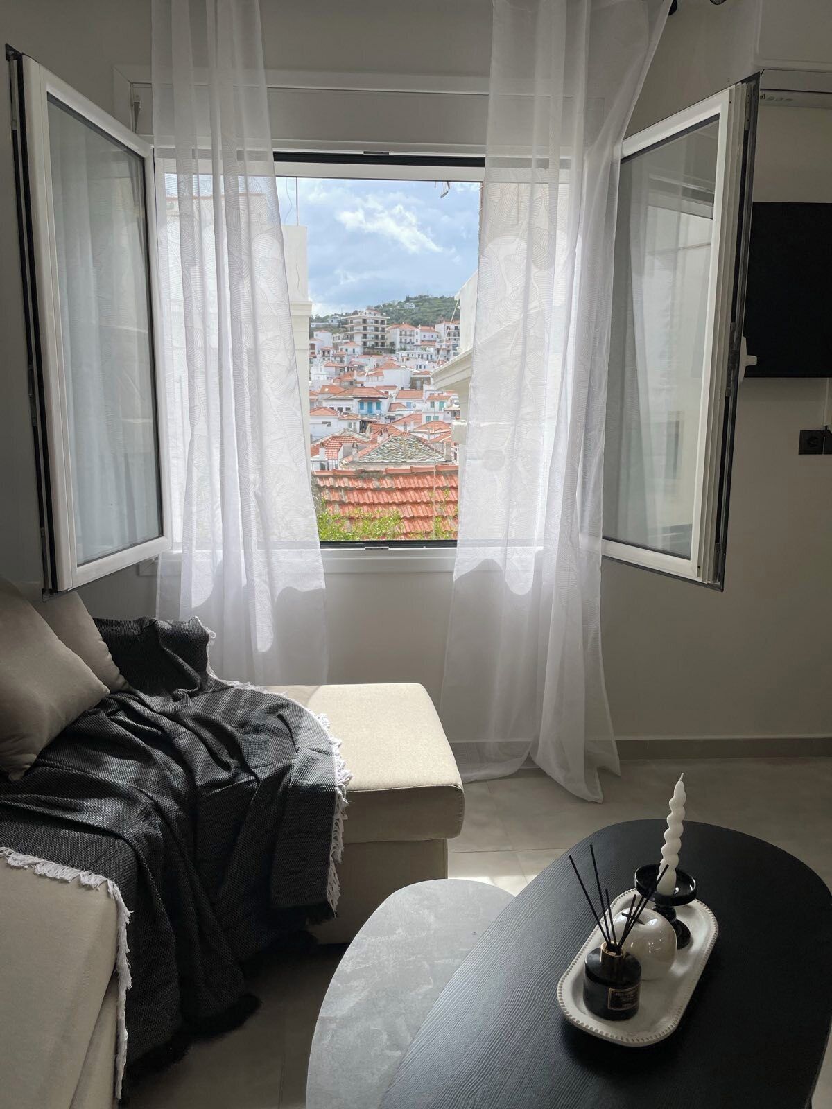 Quarto com uma cama, uma mesinha com bandeja e vista para a cidade através de uma janela com cortinas leves.