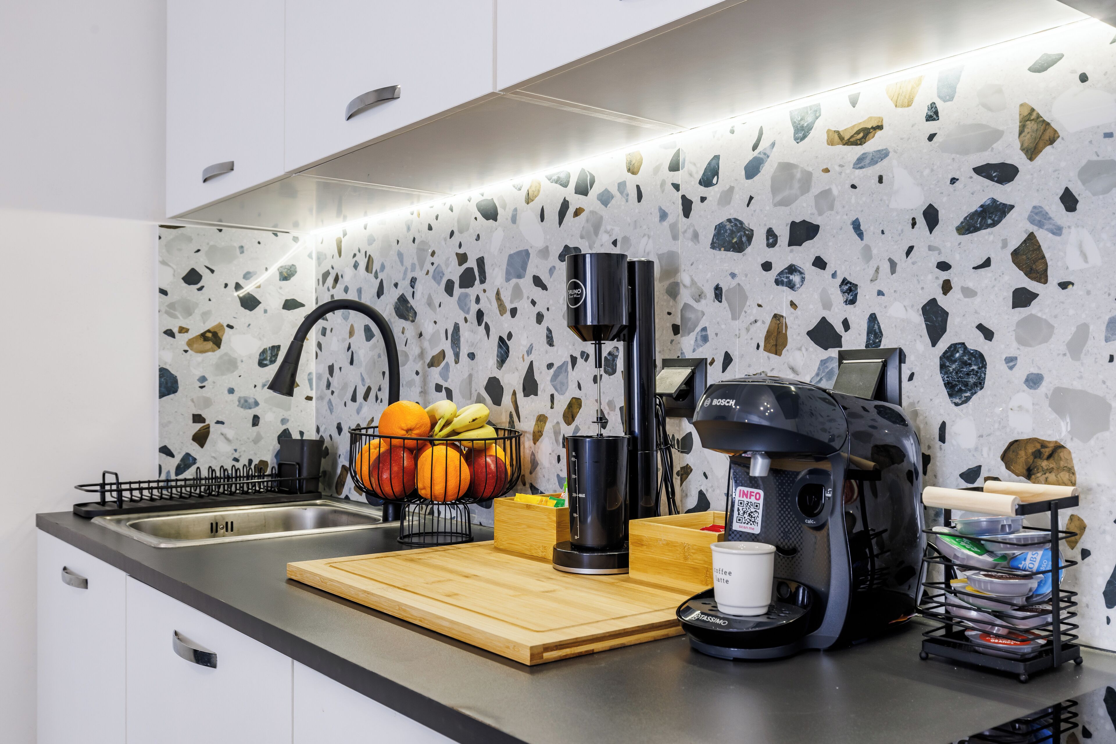 A modern kitchen with a granite backsplash, a Bosch coffee maker, a fruit bowl, and a sink.