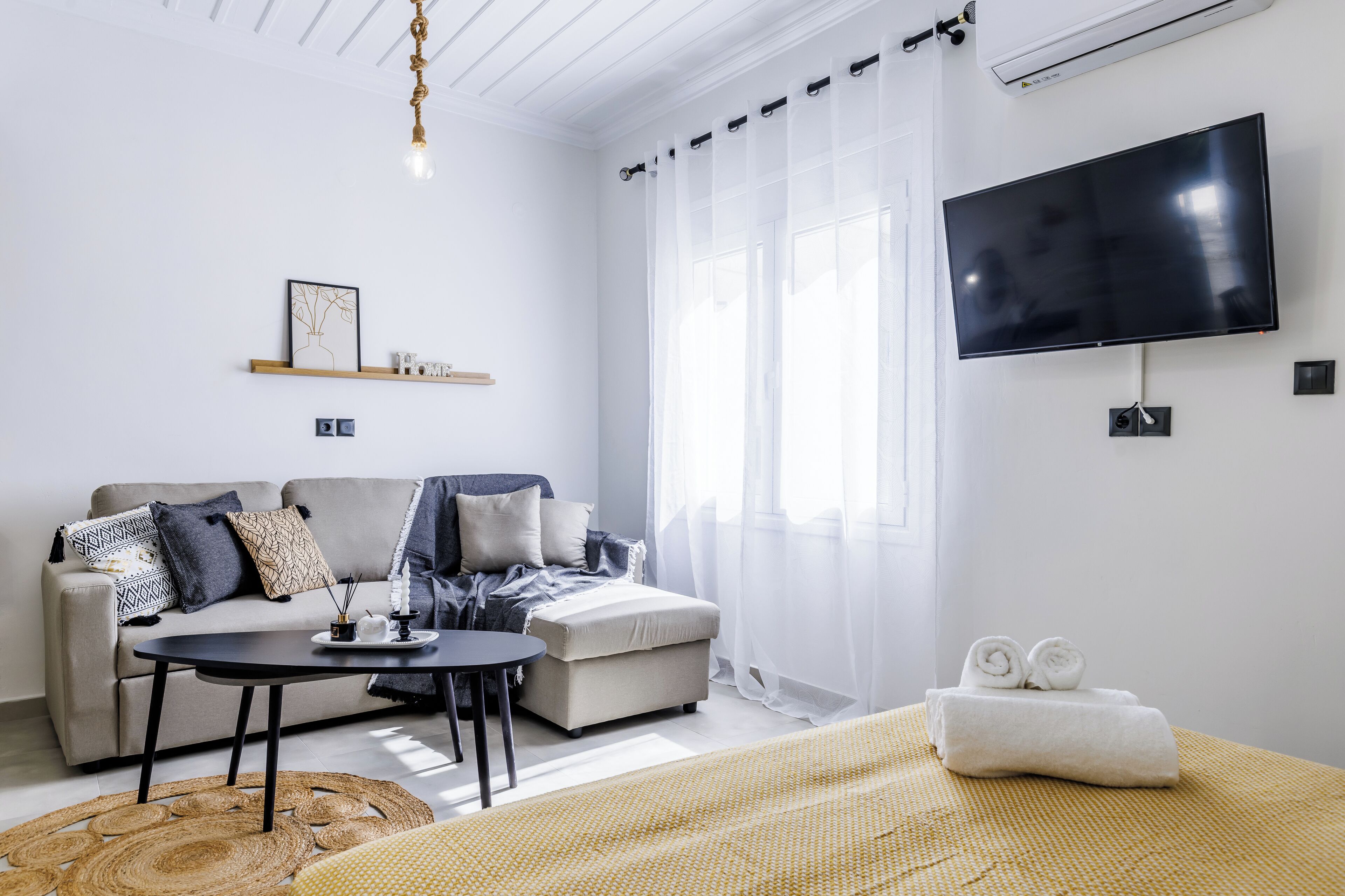 A modern living room with a sofa, a coffee table, a flat-screen TV, and a bed with rolled towels.