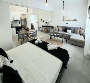 A modern hotel room with a large bed, a sofa, a dining area, and a kitchen.