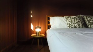 A neatly made bed with white linens and green floral pillows, a small bedside table with a lamp, and a dark wooden wall. - Obies Abode: Newly Renovated (Pittsburgh)